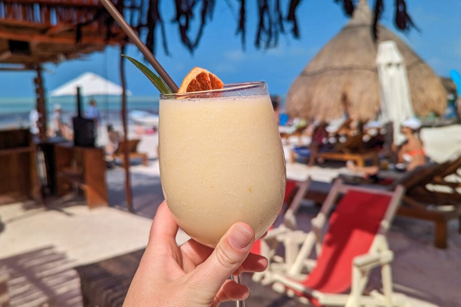 hand holding a large glass of cream coloured cocktail with an orange wedge in the top in front of a white sandy beach with red deckchairs and grass roofed palapas