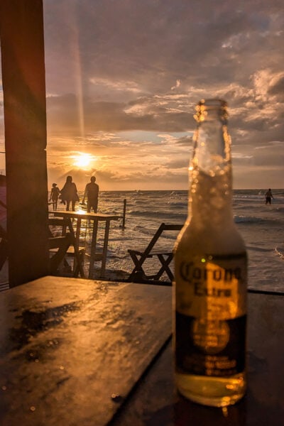 wooden table on a beach at sunset with a bottle of corona on the table and the sun setting over the sea. Best bars in Holbox