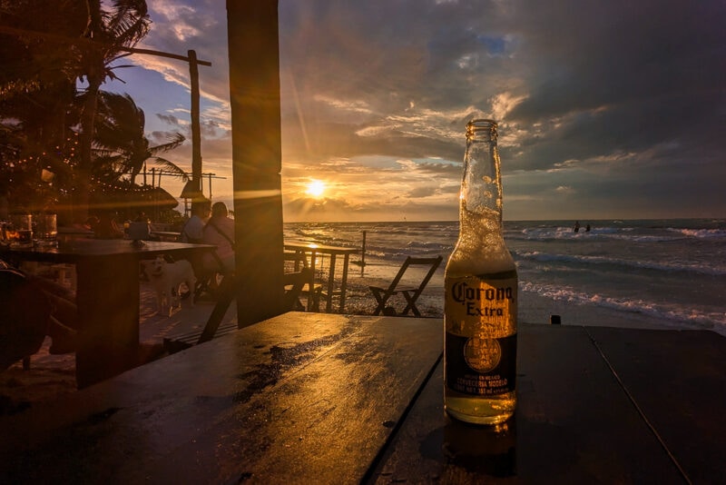 wooden table on a beach at sunset with a bottle of corona on the table and the sun setting over the sea. Best bars in Holbox