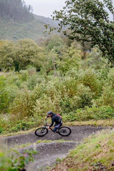 man on a mountain bike wearing a black jacket and helmet cycling downhill on a gravel path beside a green forest filled valley