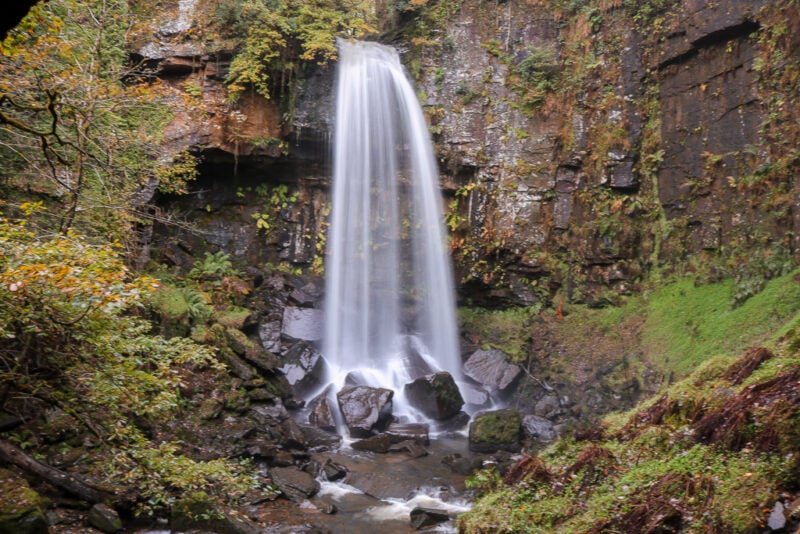large waterfall with blurred water on a grey rocky cliff face - Melincourt Waterfall. Best things to do in Neath Port Talbot South Wales UK. 