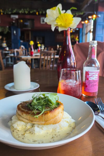 white china bowl with mashed potatoes and a circle of roast pork belly on a table next to a glass of pink lemonade with a dark restaurant interior beyond