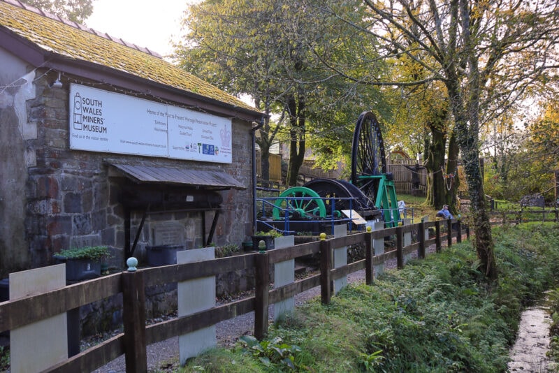 historic mining equipment, a large black metal wheel on a green frame in a forest next to a small stone building