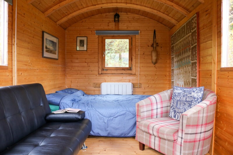 interior of a wooden shepherds hut with wood panneled walls, a double bed with blue devet, a small tartan armchair and a black leather sofa