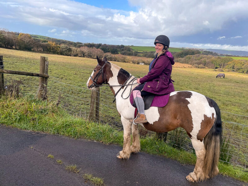emily wearing a black helmet and purple raincoat riding a white and brown horse on a country lane