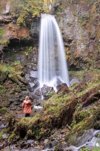 emily wearing an orange raincoat standing in front of a large waterfall with blurred water due to a long exposure. Melincourt Waterfall - Things to do in Neath Port Talbot