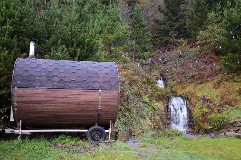 wooden outdoor sauna hut with a metal chimney next to a wooded hillside with a small waterfall. Things to do in Neath Port Talbot