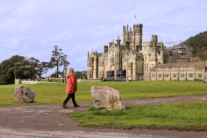 emily wearing black trousers and a long orange raincoat walking along a path towards a large stone castle with several turrets at Margam Country Park in Neath Port Talbot