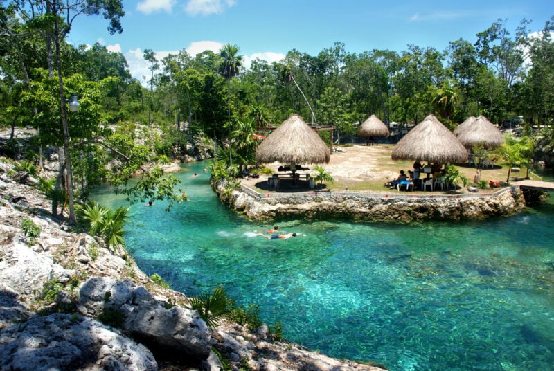 people swimming in a turquoise river with some grass roofed huts on the far side surroudned by lush green trees