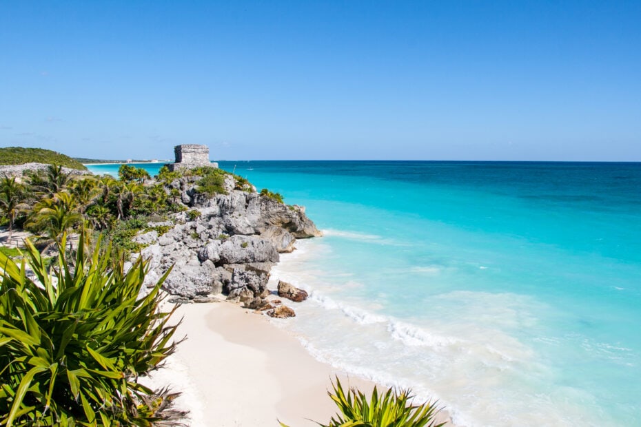 white sandy beach with turquoise sea with stone mayan ruins on a cliff in the distance