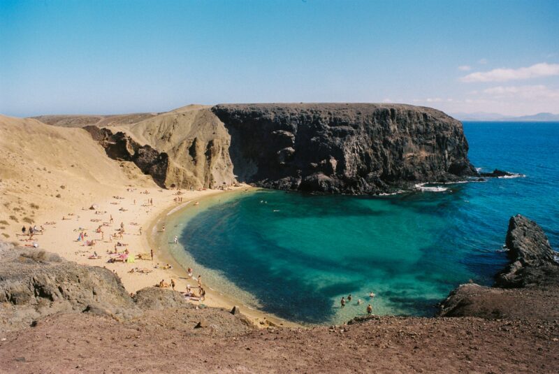 view of a semi circular bay with a sandy beach and turquoise water surrounded by brown rocky cliffs