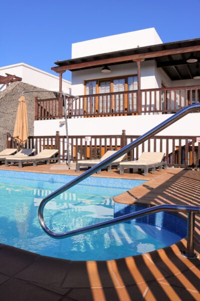 small outdoor swimming pool in front of a white painted two storey villa with wooden trim on a sunny day with blue sky