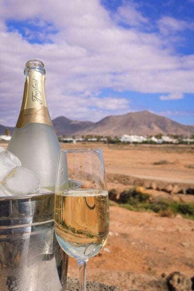 glass of cava next to a silver bucket filled with ice and a bottle of cava, with a view of a brown volcanic mountain behind