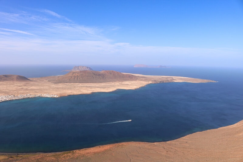view from a clifftop of the sea with a small boat passing in front of an arid, sandy island with a volcanic hill at its centre - la graciosa island seen from lanzarote in december winter