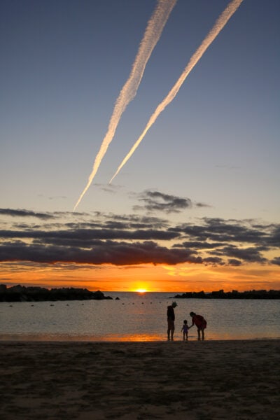silhouettes of two adults and a toddler in front of the sea at sunset with the sun just about to go behind the horizon