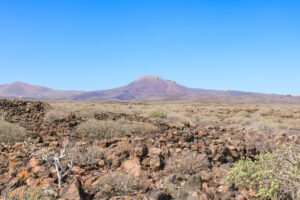 view of a distant red-brown volcano behind an aric, rocky landscape with clear blue sky above