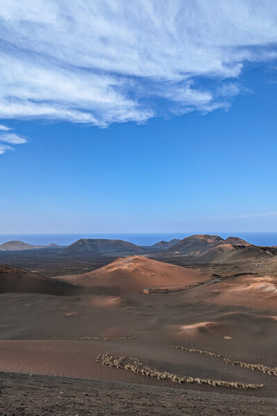 view of Timanfaya National Park with an empty, arid landscape of grey-brown rocks and volcanic cones with the sea just visible beyond