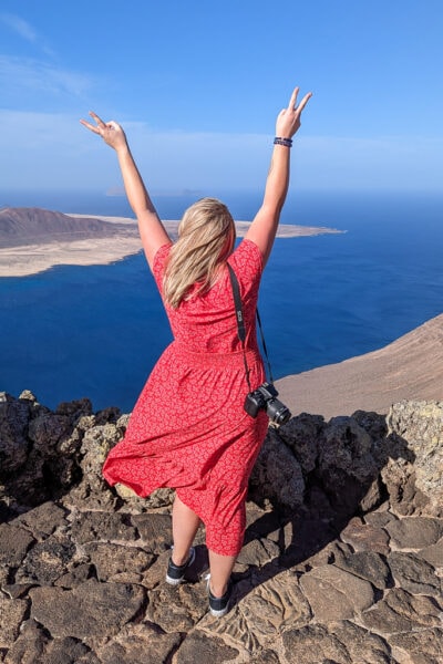 emily wearing a red midi dress with her camera on one shoulder standing in front of a viewpoint with her back to the camera and her arms in the air, the view in front of her is of the sea with an arid volcanic island