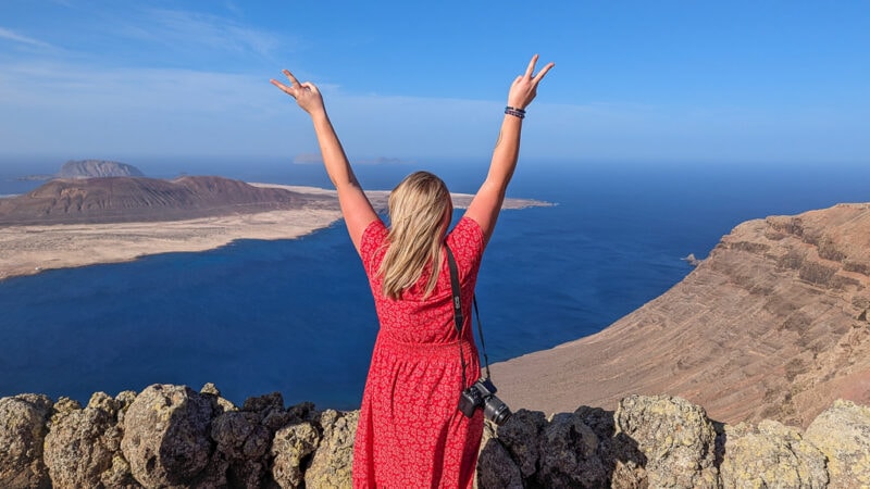 emily wearing a red midi dress with her camera on one shoulder standing in front of a viewpoint with her back to the camera and her arms in the air, the view in front of her is of the sea with an arid volcanic island. Lanzarote in Winter. 