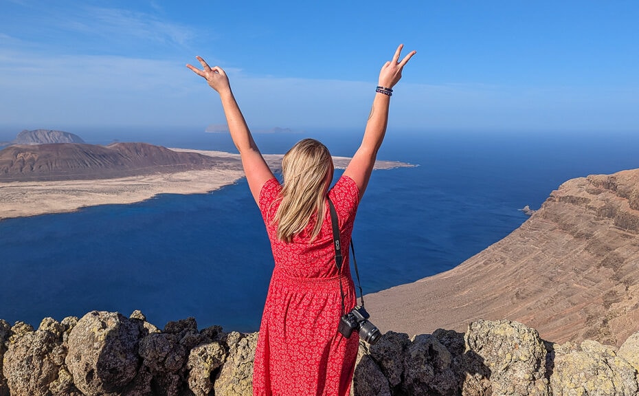 emily wearing a red midi dress with her camera on one shoulder standing in front of a viewpoint with her back to the camera and her arms in the air, the view in front of her is of the sea with an arid volcanic island. Lanzarote in Winter.