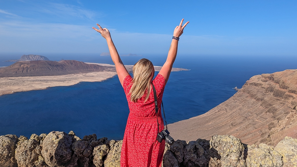 emily wearing a red midi dress with her camera on one shoulder standing in front of a viewpoint with her back to the camera and her arms in the air, the view in front of her is of the sea with an arid volcanic island. Lanzarote in Winter.