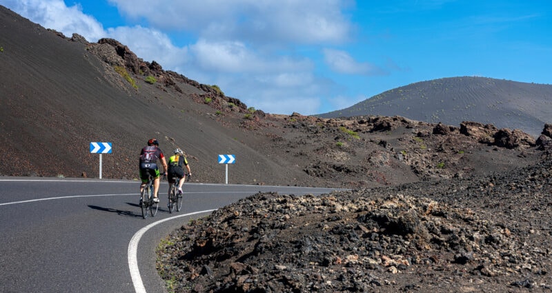 two men on mountain bikes riding on a road between grey volcanic hills 