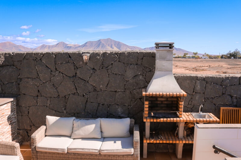outdoor seating area with cream coloured outdoor sofas and a stone and brick barbecue next to a grey stone wall with a view of brown volcanic mountains beyond