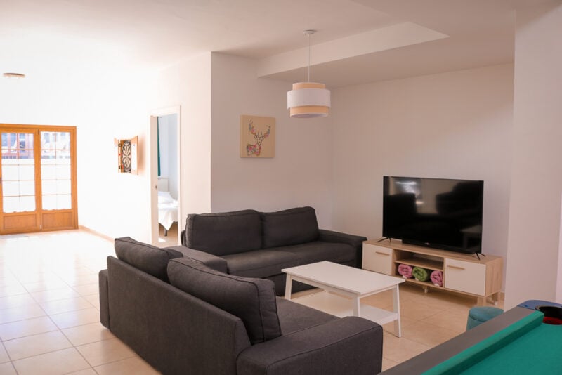 lounge with grey corner sofa arranged around a flat screen tv in a room with white walls and cream tiled floors