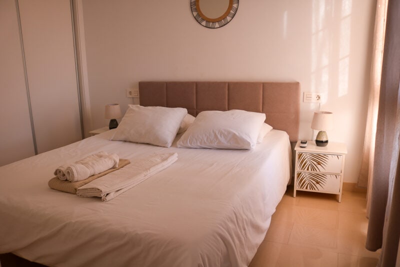 double bed with white sheets and pillows and a beige coloured head board. There is a cream coloured bedside table with gold palm leaf print next to the bed. Bedrooms in the Playa Blanca Holiday Villas in Lanzarote. 