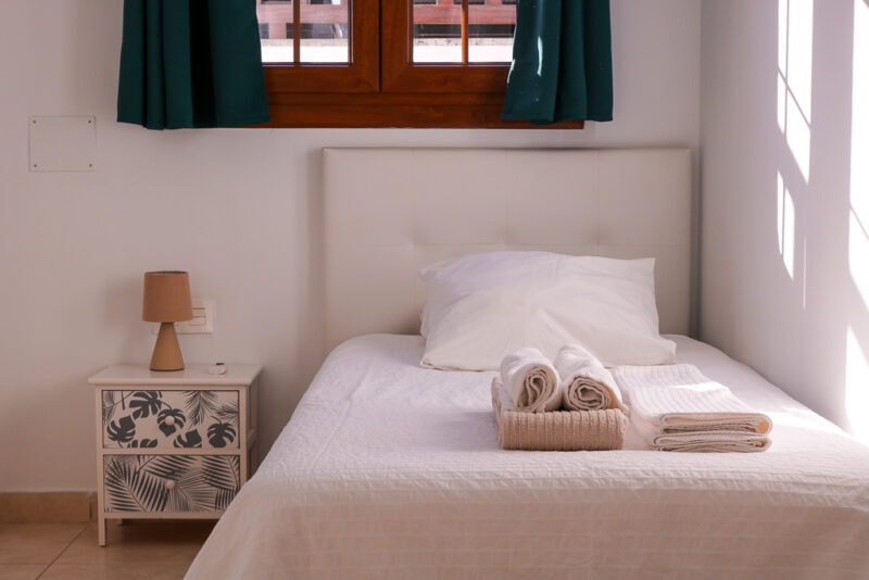 single bed with white sheets and pillow and a stack of folded towels at the end of the bed. there is a cream bedside table with blue palm leaf print next to the bed.