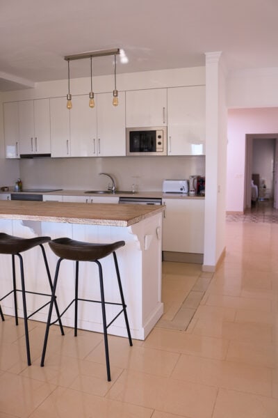 interior of a villa with cream tiled floor and white walls. There is a large wooden kichen island with two black stools and the kitchen with lots of white cupboards behind.