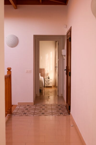 hallway with cream tiled floor and white walls, a door at the end of the hall is open with a bedroom inside and a small cream bedside table