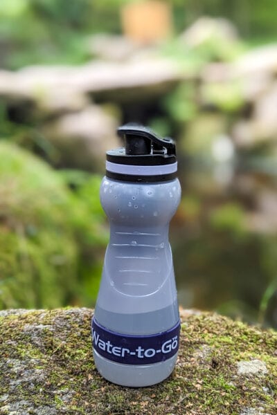 a grey bottle with purple trip and the name water-to-go printed on the side standing on a mossy rock with a river out of focus behind