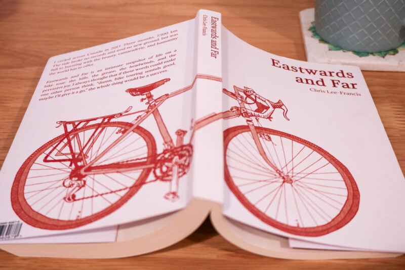white paperback book open on a table the cover has a red ink drawing on a bike on it with the name Eastwards and Far printed above