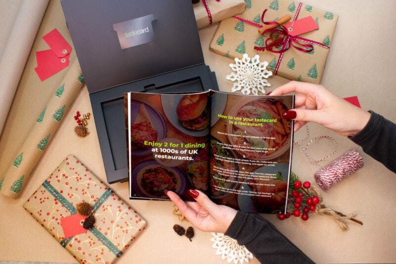 flat lay of a tabletop with wrapped christmas presents and string around an open black cardboard box with a tastecard inside and two hands in front holding an open catelogue 