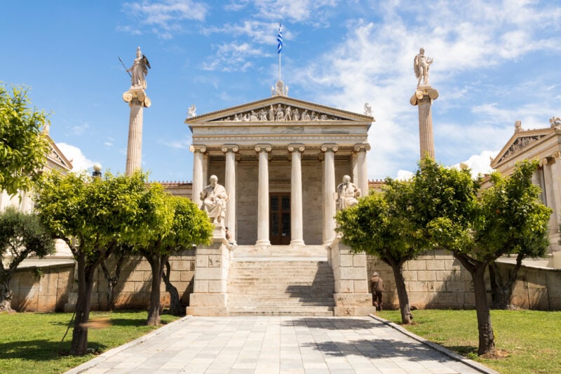 small white neoclassical style building with 6 pillars below a triangular shaped roof, there is a staircase in front and two tall white columns topped with gold statues on either side of the building