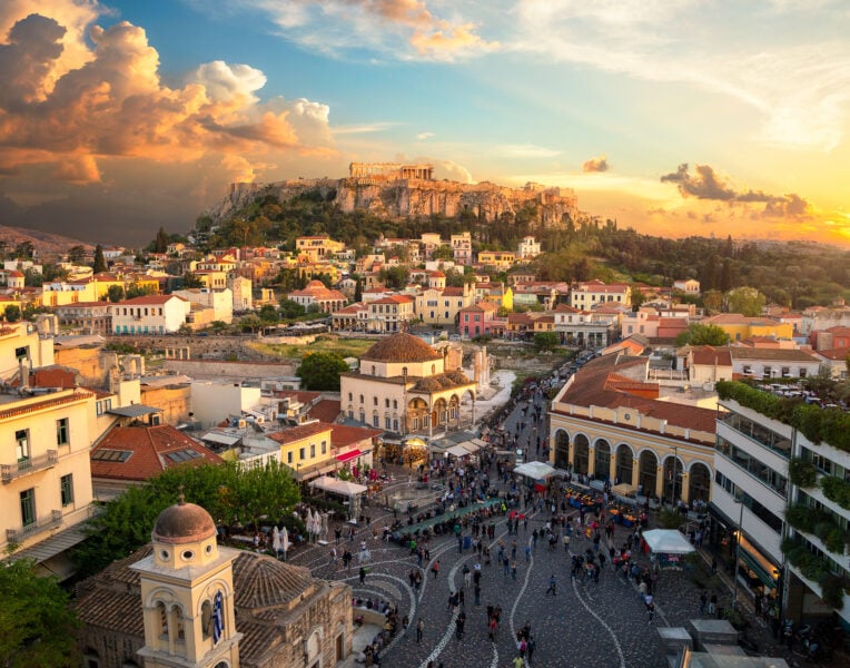 Aerial shot of the Athens city skyline taken from Monastiraki Square with a domed ottoman church with red tiled roof in the foreground on the left side of the square and a green hill in the distance with the ancient greek ruins of the acropolis on top. Unusual things to do in Athens. 