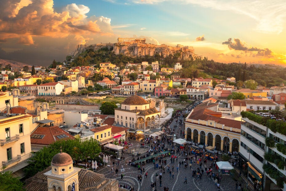 Aerial shot of the Athens city skyline taken from Monastiraki Square with a domed ottoman church with red tiled roof in the foreground on the left side of the square and a green hill in the distance with the ancient greek ruins of the acropolis on top. Unusual things to do in Athens.