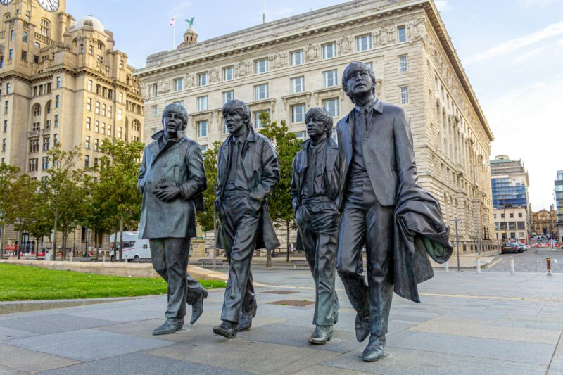 lifesize dark grey statues of 4 men in long coats walking in front of a large grey stone building - the beatles statues in Liverpool