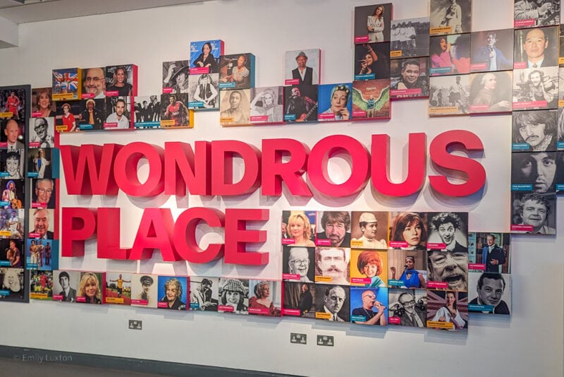 White wall in the Liverpool Museum with many portraits of different faces arranged in a cloud shape around pink letters which spell out: Wondrous Place