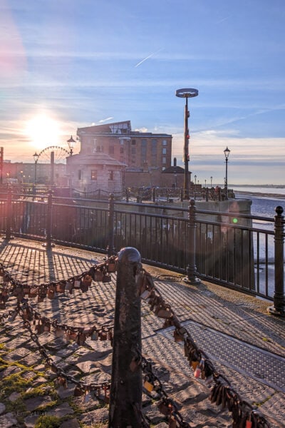Looking into a sunrise with lens flare over some red brick warehouses bext to a river and a cobbled pathway in front. there areblack iron railings in the forground with many padlocks hanging from them. Liverpool city break. 