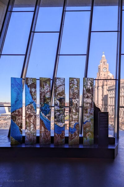 large floor to cieling window with a view of a grey stone building topped with a clock tower, in front of the window is an art work of a map painted onto glass with the light from outside coming through. Liverpool city break travel guide. 