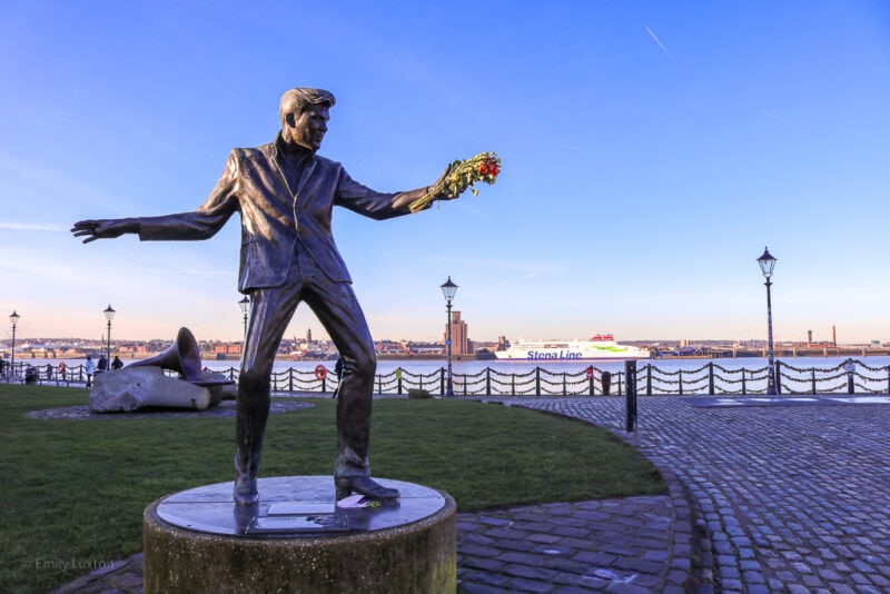 bronze statue of a man in a suit with his arms spread wide as if dancing, on a walkway in front of a river taken on a very sunny winter day with clear blue sky