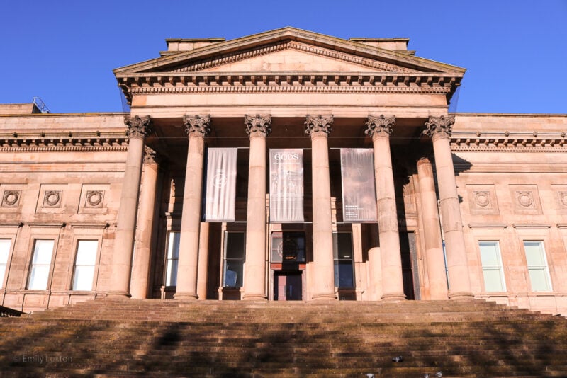 Stone steps leading up to a neoclassical building of beige stone with a triangular roof above 6 pillars on a very sunny day with clear blue sky