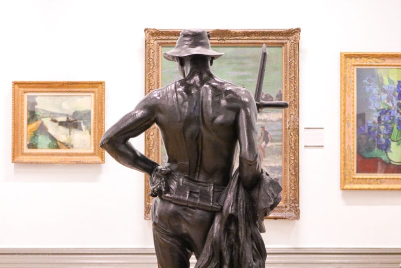 back of a bronze sculpture of a topless man wearing a floppy hat and holding a sword. The sculpture faces a white wall with three paintings of landscapes in gold frames..