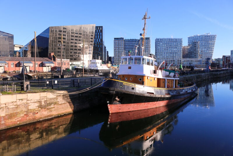 black, white and yellow fishing boat in a small dock with very still water and a skyline of tall glass skyscrapers behind. Planning a city break in liverpool.