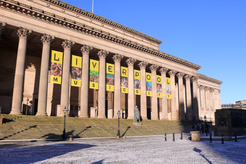 Exterior of a grand neoclassical building built from beige coloured stone with many columns. There are yellow squares with black letters in between the columnns which read: Liverpool Music City.