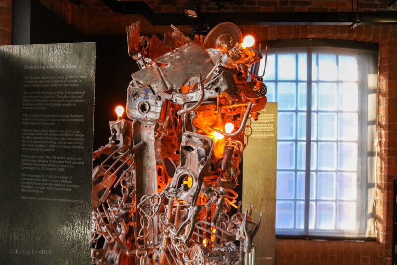 close up of a sculpture made from pieces of metal junk and light bulbs in front of an exposed red brick wall with a large window