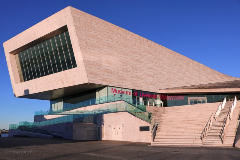 Exterior of the Museum of Liverpool a large white stone building in an angular shape with a wide stone stiarcase in front on a very sunny day with clear blue sky