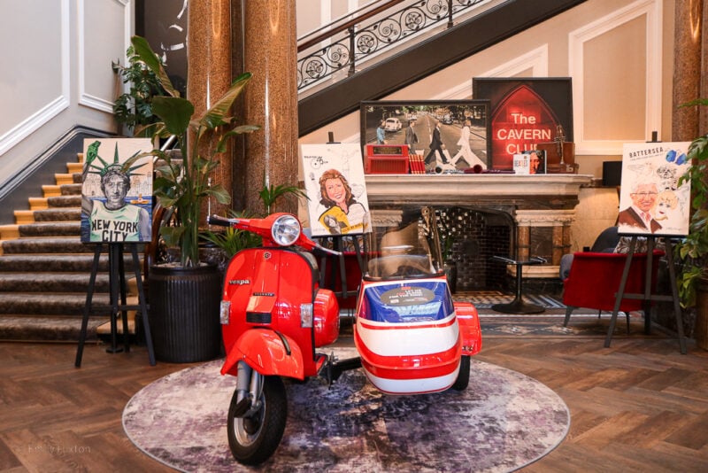 Hotel lobby with a large stone staircase behind. There is a large stone fireplace and in frotn of that a red moped with a sidecar painted in the Union Jack colours.
